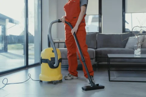 Cleaner in orange overalls vacuuming a hard floor.